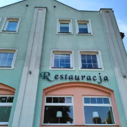 Fasada budynku restauracji z pastelową elewacją, widoczne okna z białymi ramami i napis 'Restauracja' nad wejściem