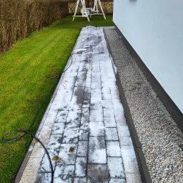 Częściowo wyczyszczona kostka brukowa na ścieżce obok białego budynku i trawnika z huśtawką w tle, widoczny wąż ciśnieniowy.