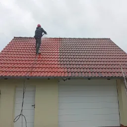 Pracownik w czerwonym kasku na drabinie czyści czerwoną dachówkę myjką ciśnieniową, widoczna różnica między czystą a brudną częścią dachu.