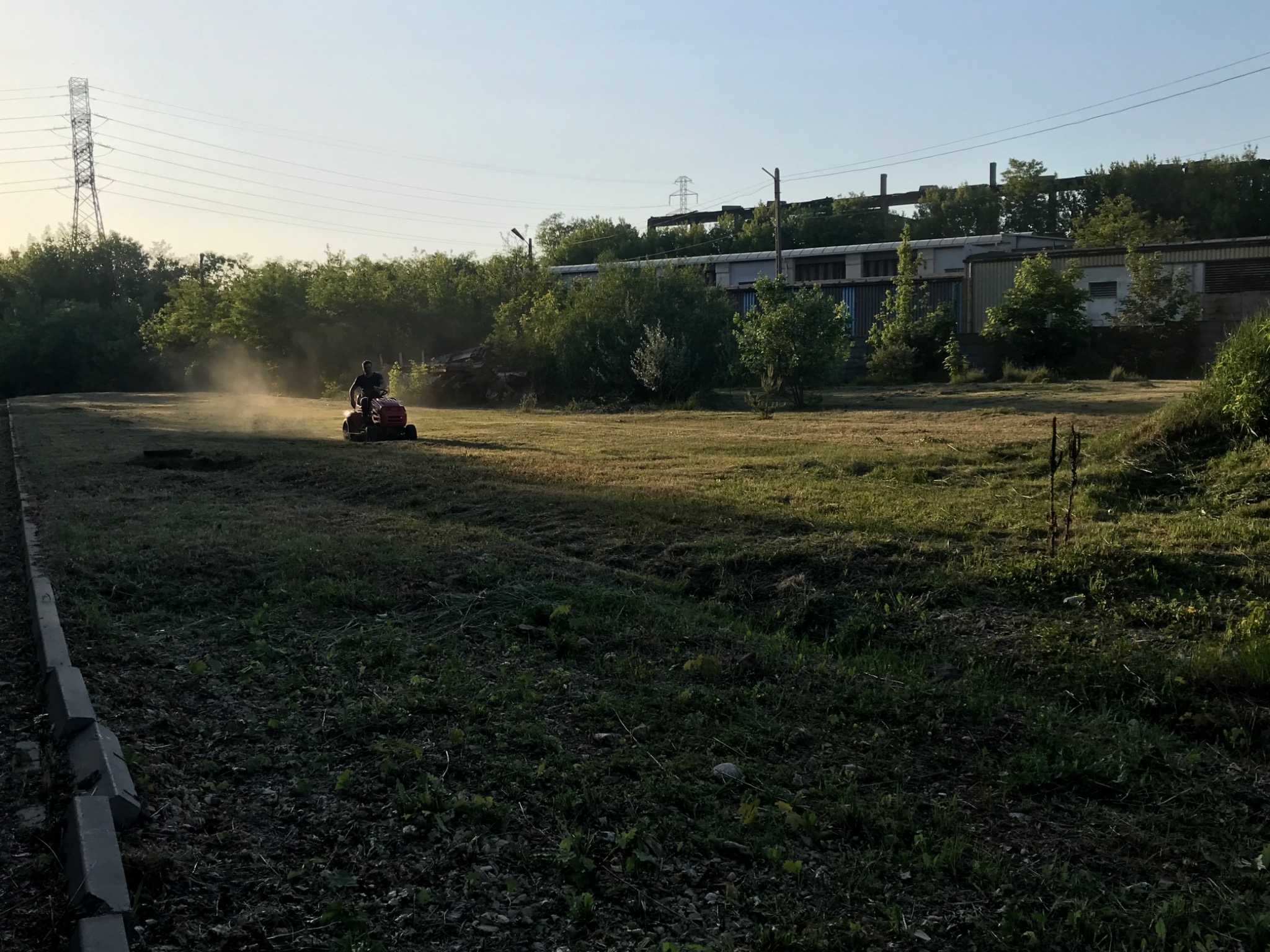 Operator kosiarką traktorkiem na rozległym trawniku, unoszący się pył, w tle budynki przemysłowe i słup wysokiego napięcia.