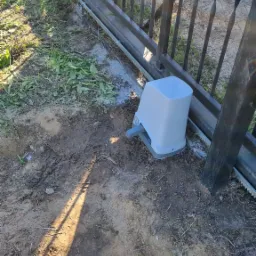 Szary napęd elektryczny do bramy przesuwnej, zamontowany przy metalowej konstrukcji bramy z zębatką, widoczny na ziemi z fragmentami trawy.