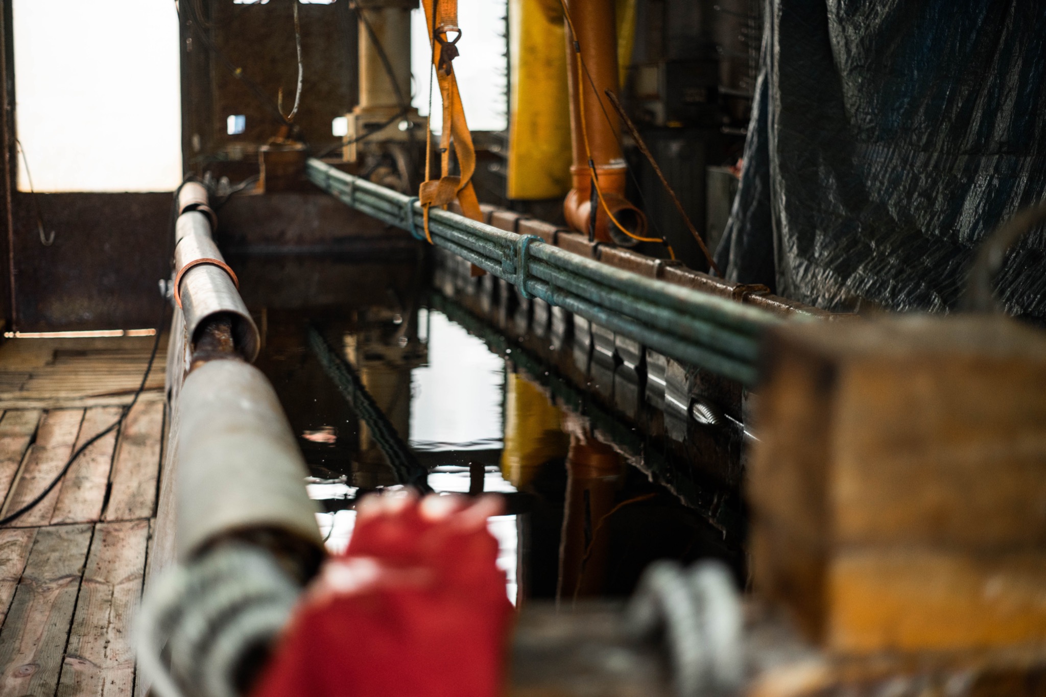 Wnętrze warsztatu z metalowymi rurami zawieszonymi nad zbiornikiem z wodą, widoczne odbicia i elementy konstrukcyjne w tle.