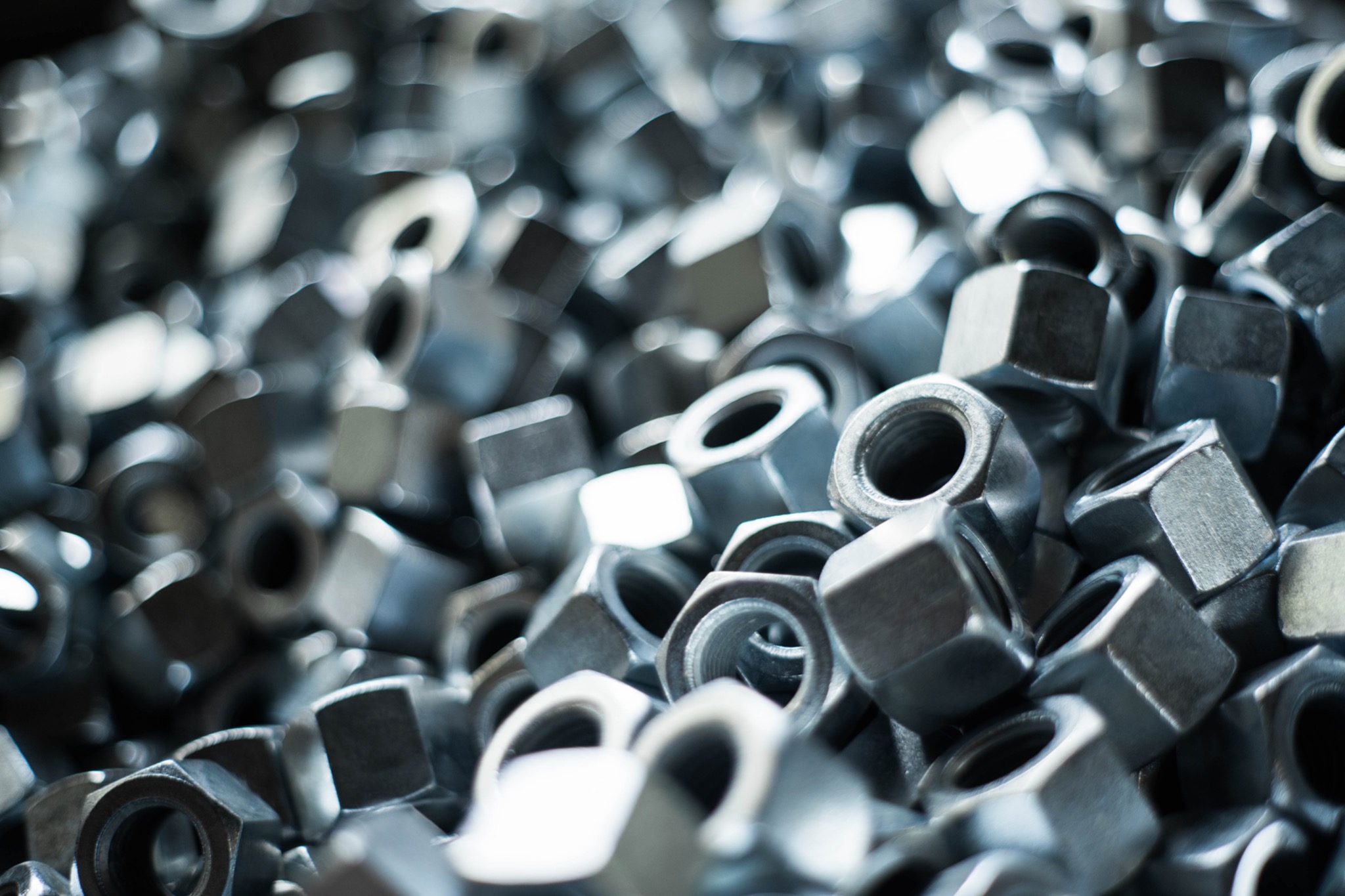 Stalowe nakrętki sześciokątne ułożone chaotycznie, wypełniające kadr, z widocznymi gwintami wewnętrznymi i delikatnymi refleksami światła.