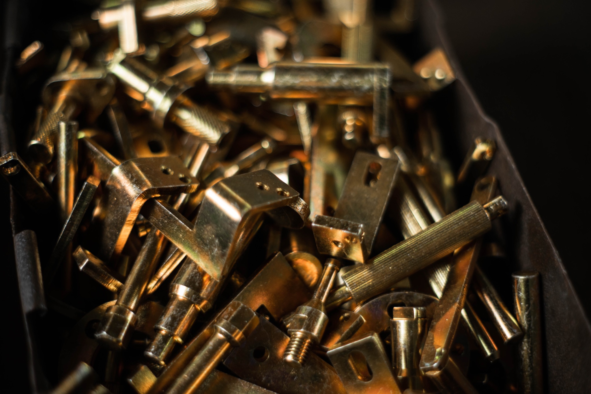 Zestaw pozłacanych, metalowych elementów konstrukcyjnych o różnych kształtach, w tym kątowniki, pręty gwintowane i tuleje, zgromadzonych w ciemnym pojemniku.