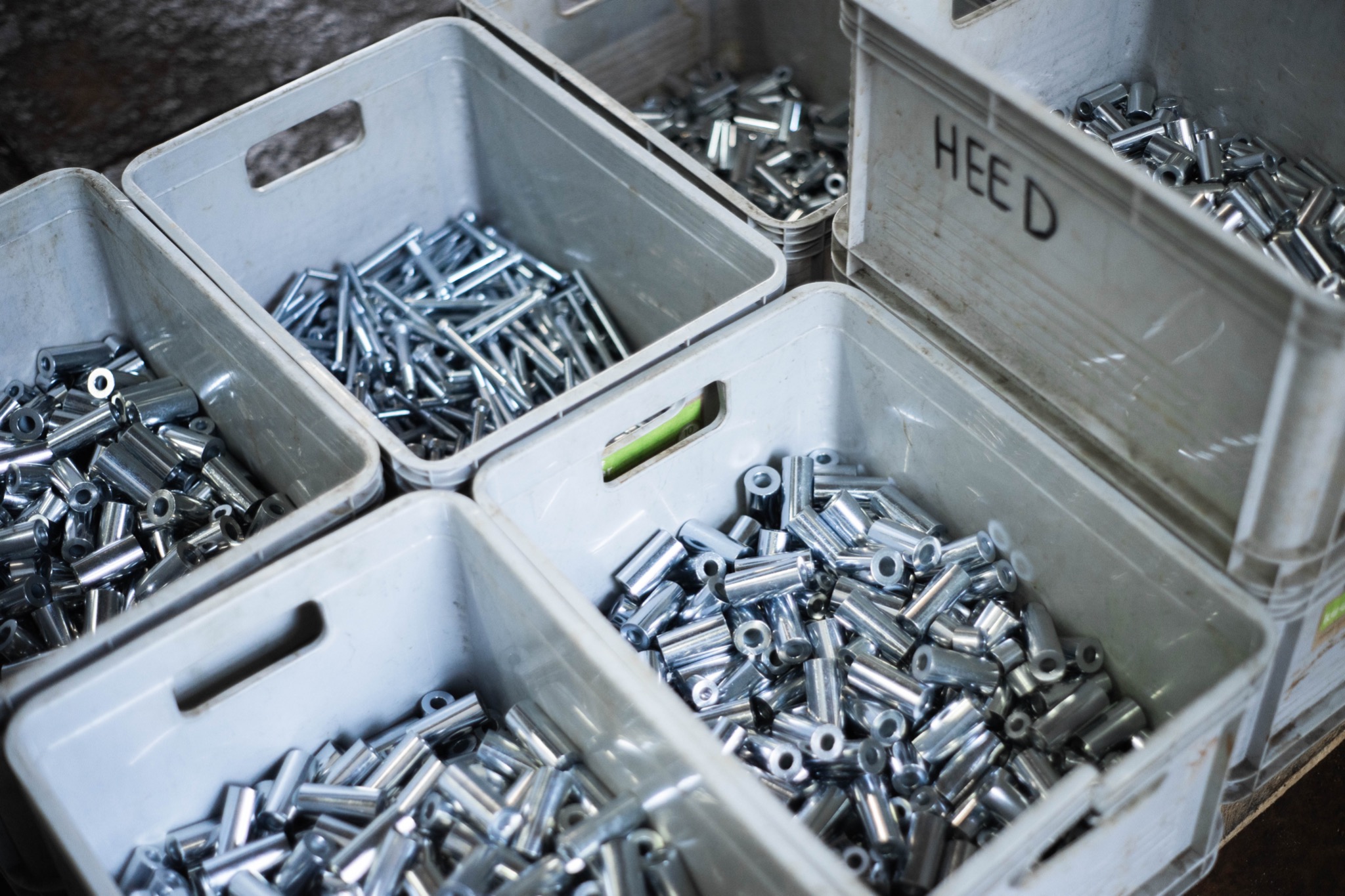 Srebrne, metalowe tuleje i pręty w szarych plastikowych pojemnikach, jeden z napisem 'HEED'.