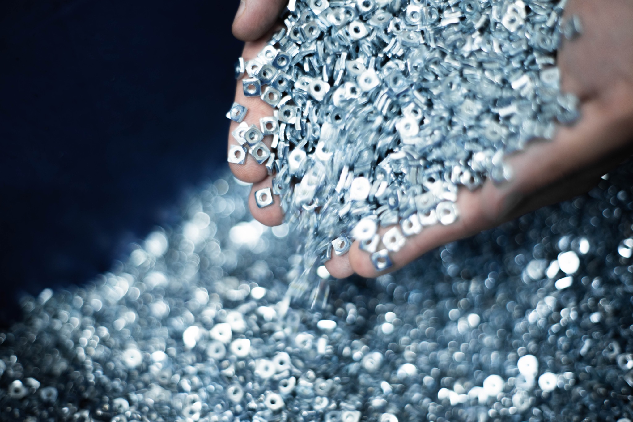 Garść srebrnych, kwadratowych nakrętek z otworem pośrodku, przesypujących się przez palce na niebieskim tle.
