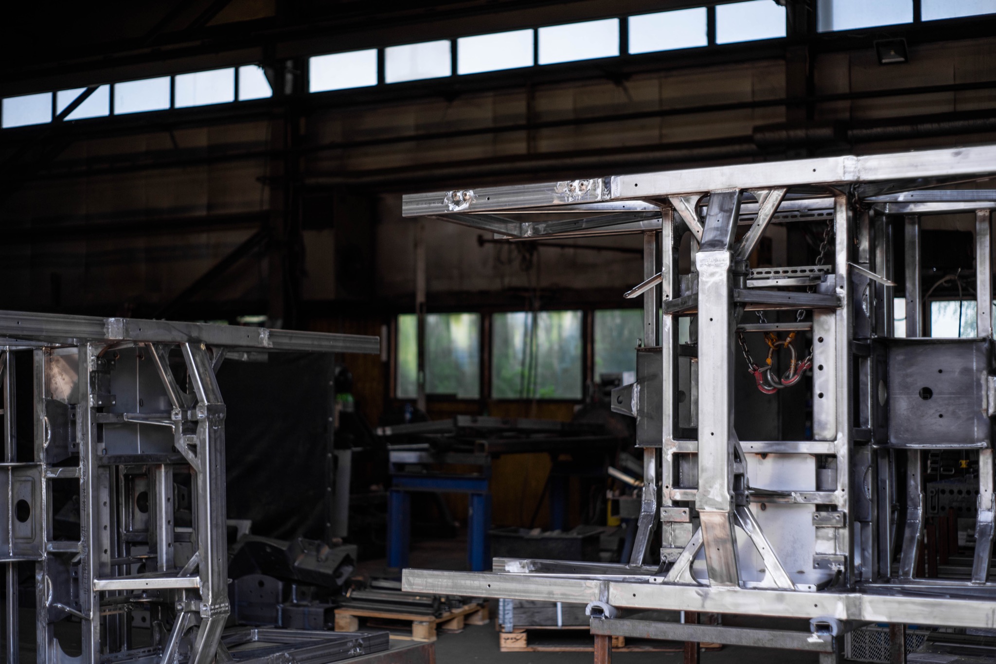 Metalowa konstrukcja szkieletowa w warsztacie, elementy spawane, widoczne spawy i przygotowane otwory, surowe wykończenie, oświetlenie naturalne z okien.