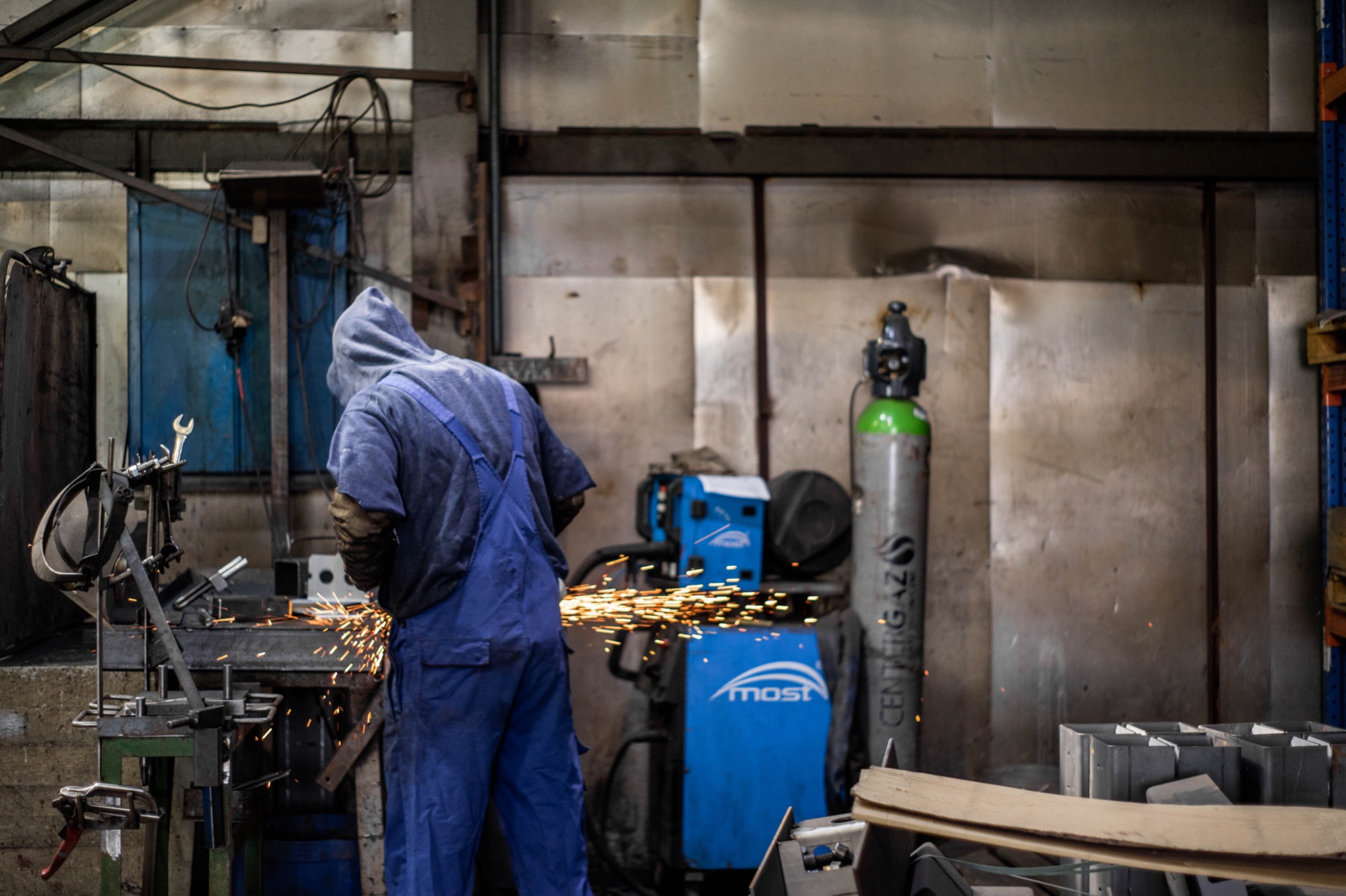 Pracownik w niebieskim kombinezonie i kapturze, odwrócony tyłem, wykonuje obróbkę metalu iskrzącą się iskrami, na tle warsztatu z butlą gazową i urządzeniem spawalniczym marki MOST.