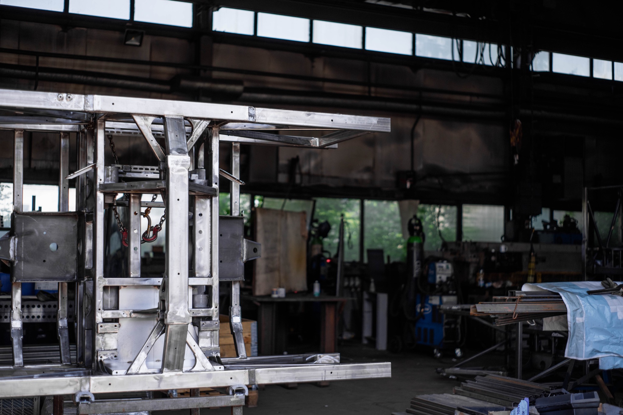 Metalowa konstrukcja ramy w warsztacie, z widocznymi spawami, łańcuchem z hakiem i elementami wyposażenia warsztatu w tle.