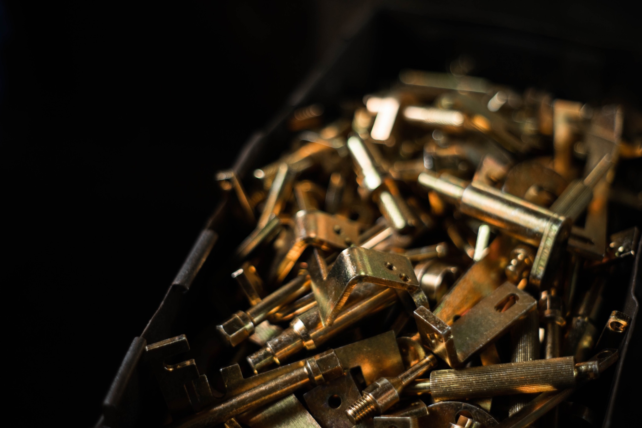 Stalowe, złote elementy z gwintami i otworami, układane w czarnym pojemniku, w ciemnym otoczeniu.