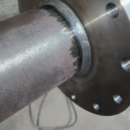 Spoina łącząca rurę z kołnierzem, widoczna tęczowa barwa spoiny. Szlifowany kołnierz z otworami na śruby. Widoczny fragment przewodu spawalniczego.