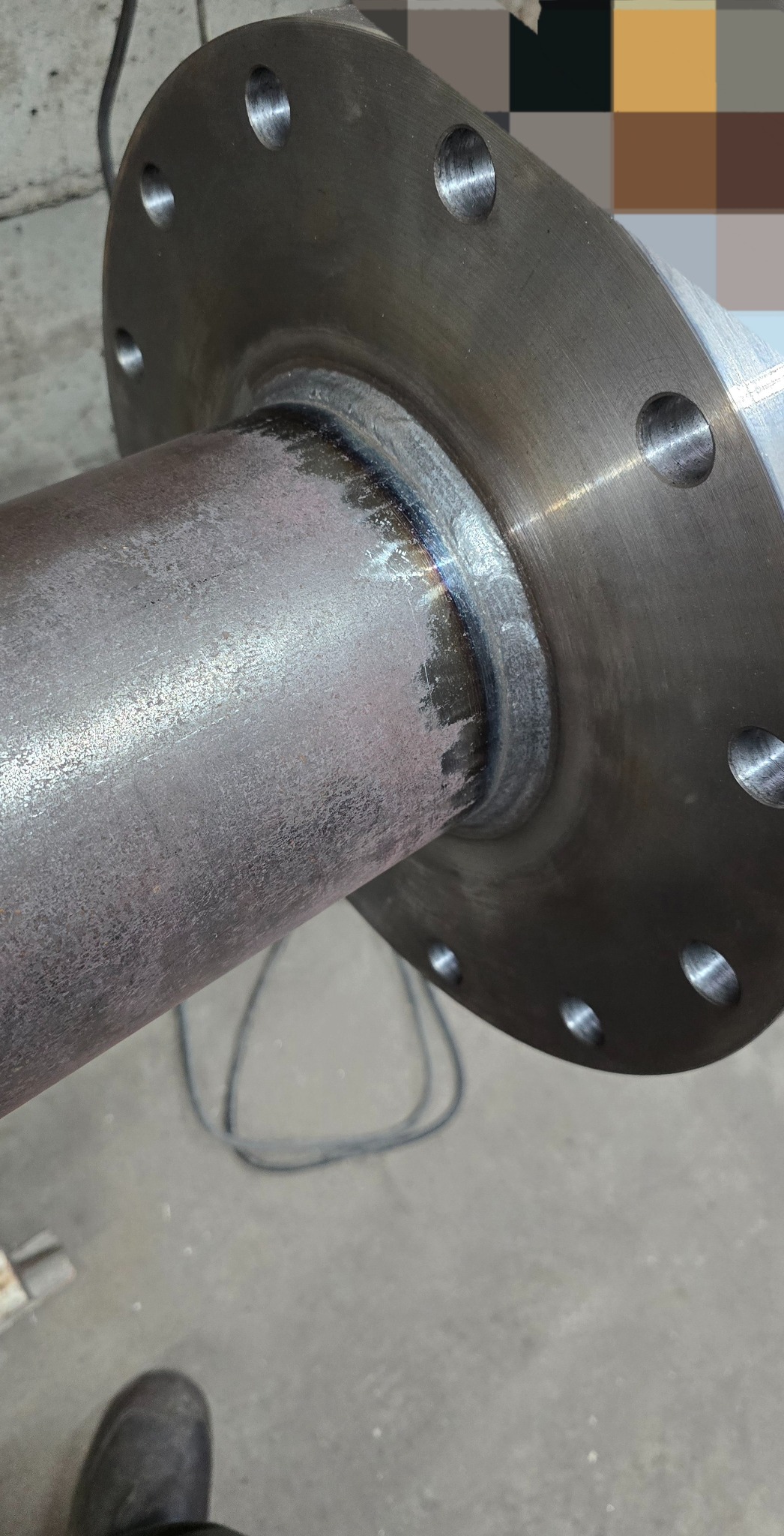 Spoina łącząca rurę z kołnierzem, widoczna tęczowa barwa spoiny. Szlifowany kołnierz z otworami na śruby. Widoczny fragment przewodu spawalniczego.