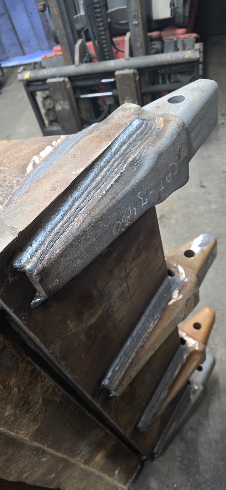 Detal spawania z bliska na zębie łyżki koparki z napisem 'CAT-J 450', widoczny w tle wózek widłowy.