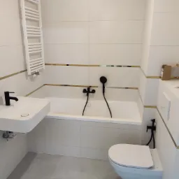 Nowoczesna łazienka w bieli z czarną armaturą: umywalka, bateria wannowa i bidetta, wykończona płytkami z delikatnym złotym akcentem.