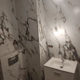 Wykończona łazienka z białymi płytkami imitującymi marmur, wisząca toaleta i szafka z umywalką, widoczne instalacje pod baterię.