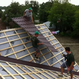 Trzech dekarzy w zielonych czapkach montuje brązowe panele dachowe na drewnianej konstrukcji dachu domu jednorodzinnego, widok z góry na plac budowy.