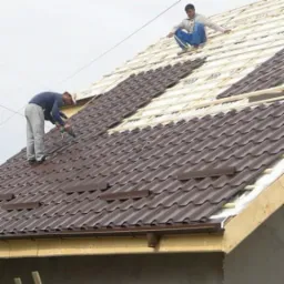 Dwóch dekarzy montuje brązowe blaszane panele na drewnianej konstrukcji dachu, jeden przykręca panele, a drugi siedzi na górze i mierzy odległość.