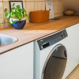 BlackForest Adrian Bonowski - Jasny blat drewniany z wbudowanym zlewem i pralką Haier. Na blacie roślina w niebieskiej doniczce, brązowe pudełko i dekoracje. Płytki na ścianie w tle.