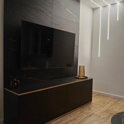 BlackForest Adrian Bonowski - Czarna szafka RTV z marmurowym panelem ściennym i złotymi akcentami, nowoczesne oświetlenie liniowe, drewniana podłoga. Elegancki wystrój salonu.