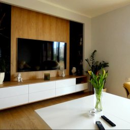 BlackForest Adrian Bonowski - Nowoczesna aranżacja salonu z drewnianą zabudową TV, białymi szafkami i czarnymi akcentami. Na pierwszym planie biały stół z tulipanami i pilotami.