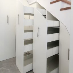 BlackForest Adrian Bonowski - Biała zabudowa pod schodami z wysuwanymi szufladami i szafkami. Minimalistyczny design, szare płytki podłogowe. Funkcjonalne wykorzystanie przestrzeni.