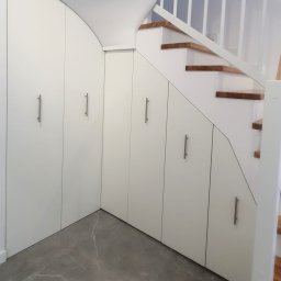 BlackForest Adrian Bonowski - Biała zabudowa pod schodami z szufladami i szafkami z podłużnymi uchwytami. Szare płytki podłogowe, drewniane schody z białą balustradą.