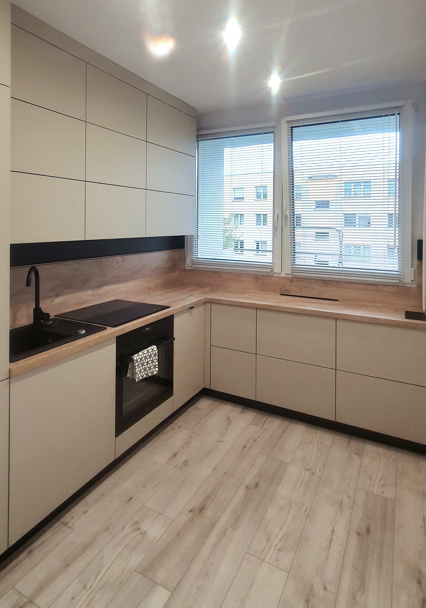 Jasna kuchnia z drewnianym blatem i czarnymi akcentami, widok na okno z żaluzjami. Minimalistyczny design, nowoczesne wykończenie, oświetlenie sufitowe.