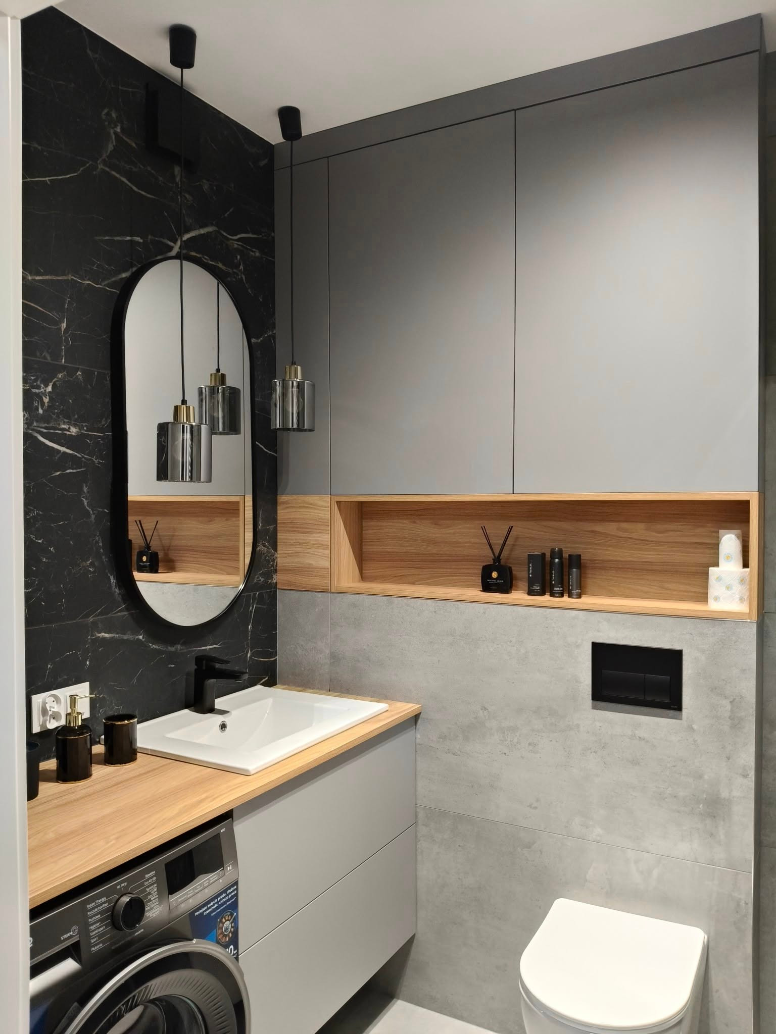 Nowoczesna łazienka z szarą zabudową, blatem drewnianym nad pralką i czarnym marmurem na ścianie. Owalne lustro i wiszące lampy dodają elegancji.