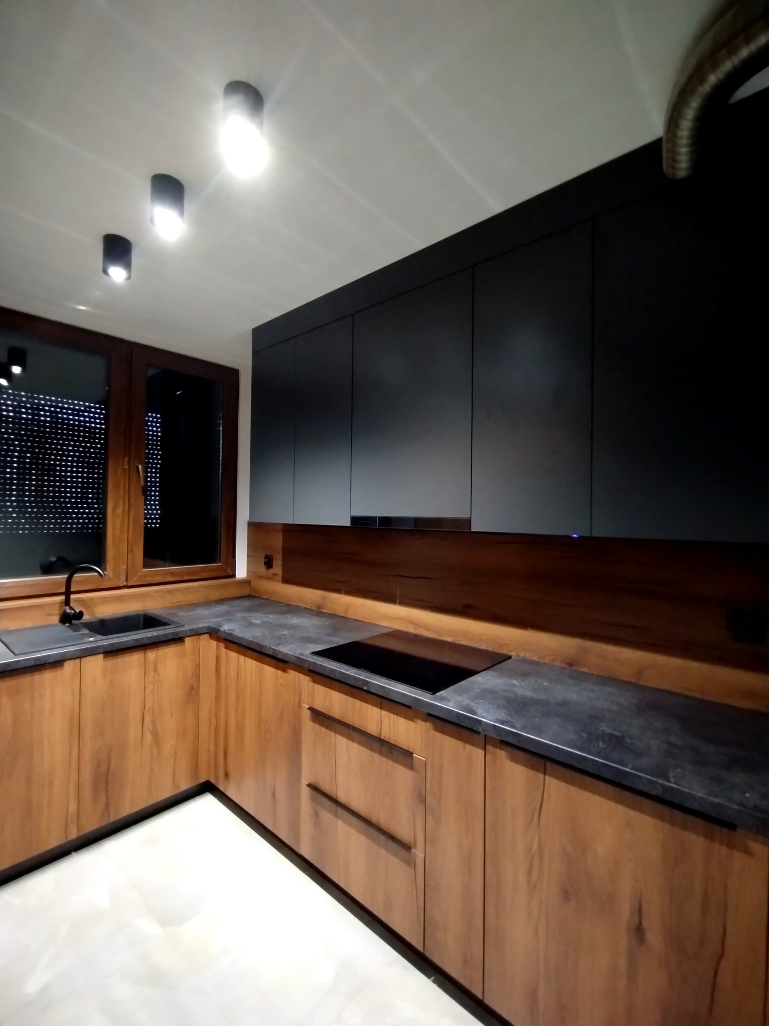 Nowoczesna kuchnia z drewnianymi szafkami dolnymi i czarnymi górnymi, czarny blat, okno z ciemną ramą i punktowe oświetlenie sufitowe.