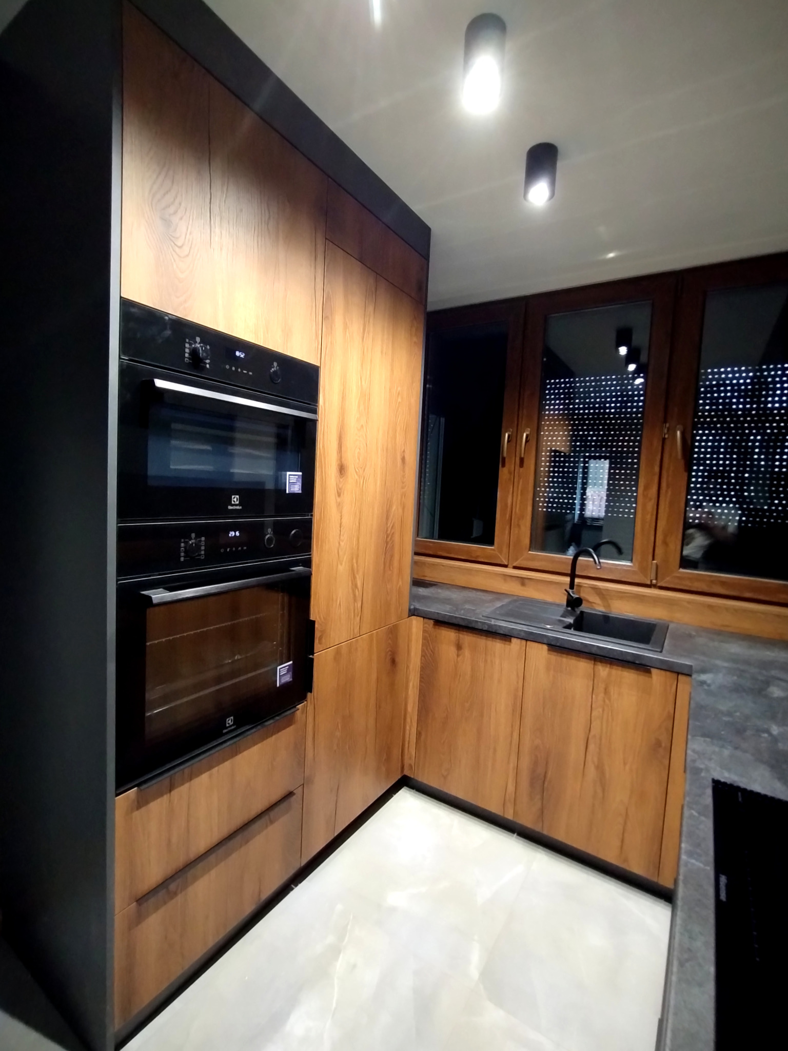 Nowoczesna kuchnia z drewnianymi frontami, czarnym blatem i sprzętem AGD Electrolux. Ujęcie z niskiego kąta podkreśla głębię i fakturę materiałów.