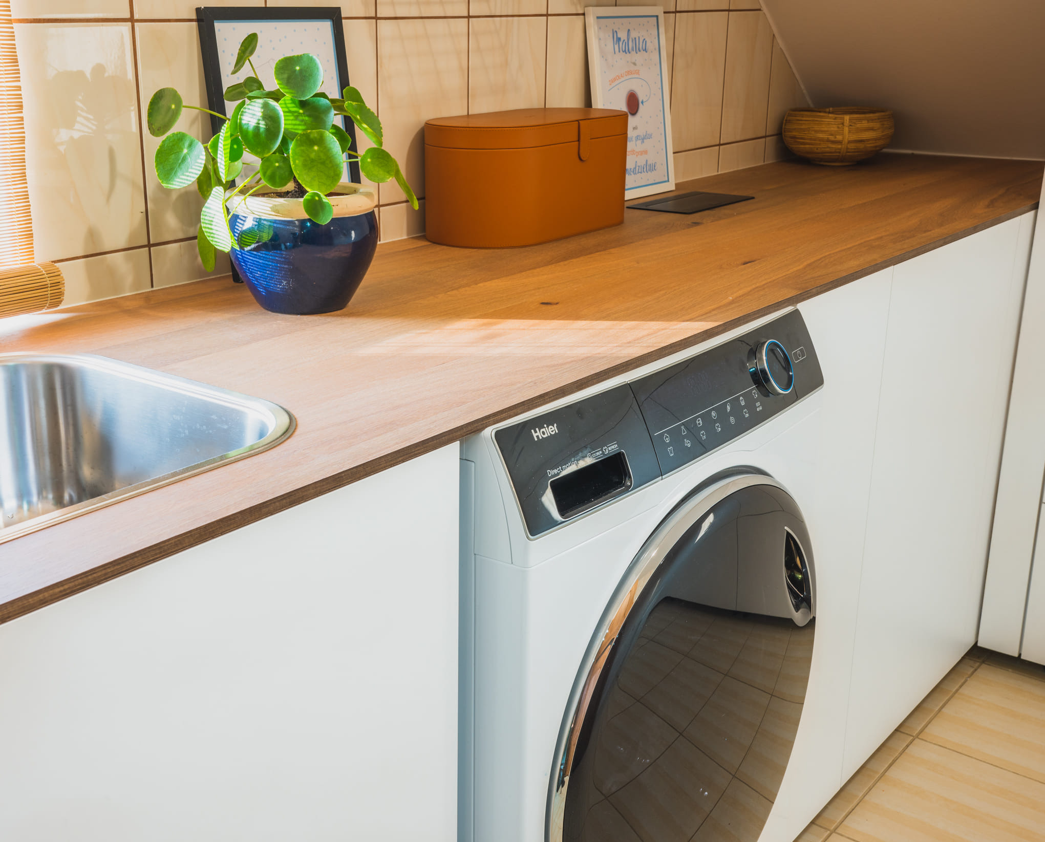 Jasny blat drewniany z wbudowanym zlewem i pralką Haier. Na blacie roślina w niebieskiej doniczce, brązowe pudełko i dekoracje. Płytki na ścianie w tle.