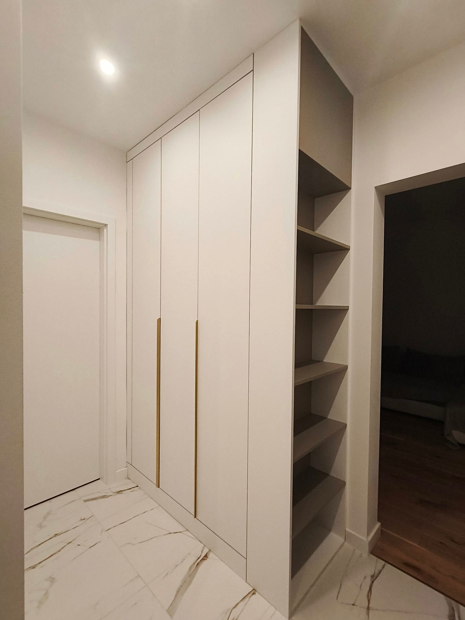 Biała szafa wnękowa z złotymi uchwytami i otwartymi półkami w przedpokoju z marmurową podłogą. Minimalistyczny design, oświetlenie sufitowe.
