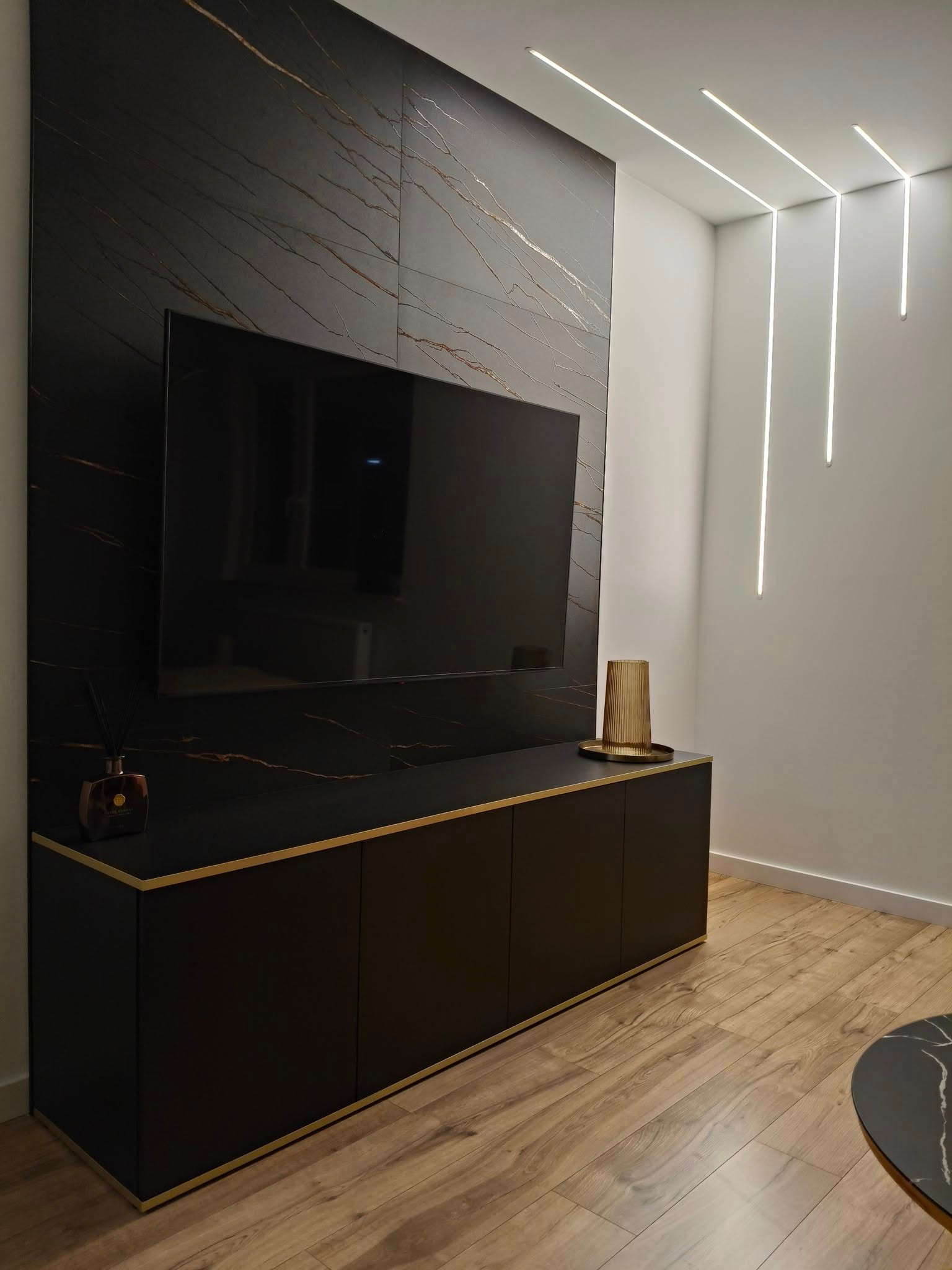 Czarna szafka RTV z marmurowym panelem ściennym i złotymi akcentami, nowoczesne oświetlenie liniowe, drewniana podłoga. Elegancki wystrój salonu.