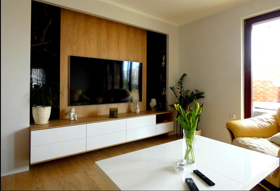 Nowoczesna aranżacja salonu z drewnianą zabudową TV, białymi szafkami i czarnymi akcentami. Na pierwszym planie biały stół z tulipanami i pilotami.