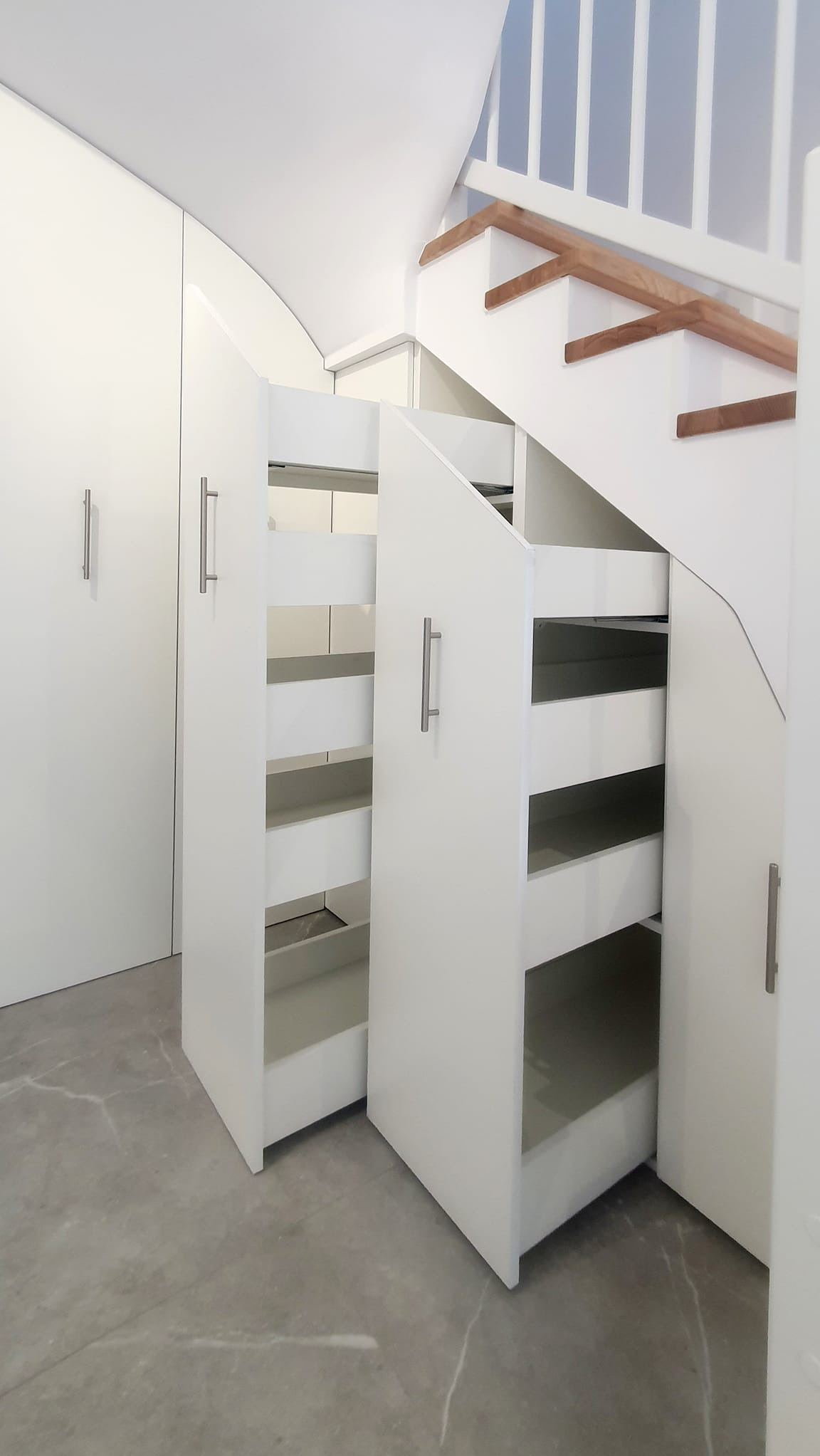 Biała zabudowa pod schodami z wysuwanymi szufladami i szafkami. Minimalistyczny design, szare płytki podłogowe. Funkcjonalne wykorzystanie przestrzeni.
