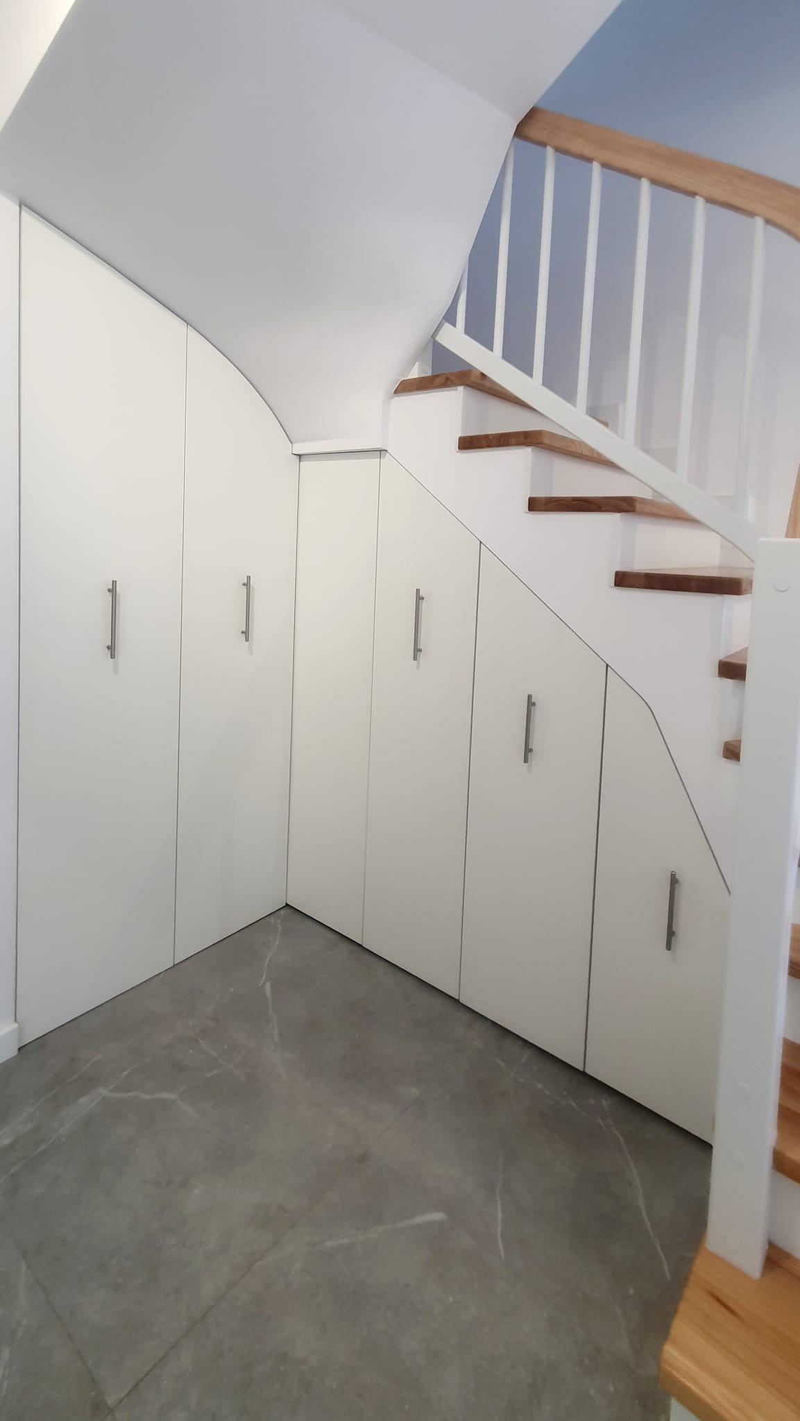 Biała zabudowa pod schodami z szufladami i szafkami z podłużnymi uchwytami. Szare płytki podłogowe, drewniane schody z białą balustradą.