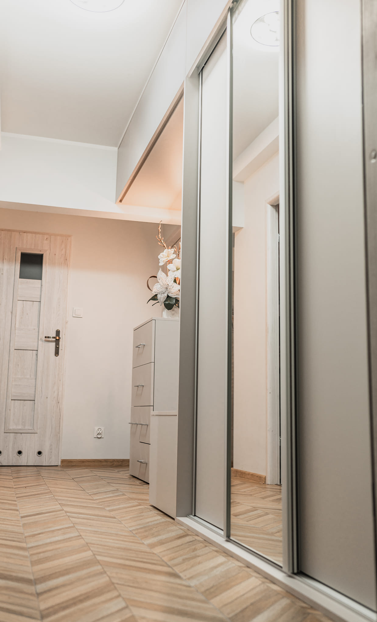 Jasne wnętrze z szafą przesuwną z lustrami, komodą z kwiatami i drewnianą podłogą ułożoną w jodełkę. Widoczne drzwi wejściowe w tle. Minimalistyczny design.