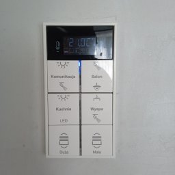 AUTOMATYKA-INSTALACJE ELEKTRYCZNE - PROJEKTY ZENON REMIŚKO - Panel sterowania inteligentnego domu z wyświetlaczem temperatury i ikonami oświetlenia dla różnych pomieszczeń, w tym kuchni, salonu i wyspy. Widoczne przyciski sterowania roletami.