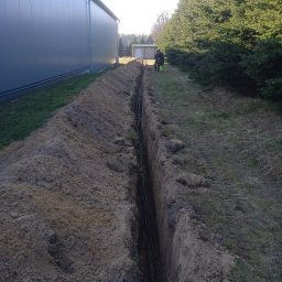 AUTOMATYKA-INSTALACJE ELEKTRYCZNE - PROJEKTY ZENON REMIŚKO - Wykop z ułożonymi kablami elektrycznymi w ziemi, przygotowany do zasypania. Widoczny budynek i rząd drzew w tle. Prace instalacyjne w toku.