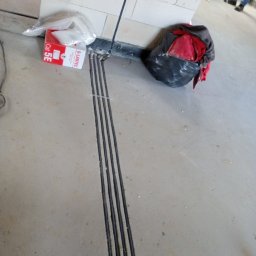 AUTOMATYKA-INSTALACJE ELEKTRYCZNE - PROJEKTY ZENON REMIŚKO - Wykute bruzdy w posadzce pod kable elektryczne, obok puszka instalacyjna i worek z odpadami. Widoczne fragmenty instalacji w ścianie.