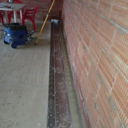 AUTOMATYKA-INSTALACJE ELEKTRYCZNE - PROJEKTY ZENON REMIŚKO - Metalowe korytka kablowe ułożone wzdłuż ceglanej ściany, przygotowane do montażu. W tle widoczny odkurzacz przemysłowy i krzesła.