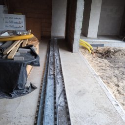 AUTOMATYKA-INSTALACJE ELEKTRYCZNE - PROJEKTY ZENON REMIŚKO - Metalowe korytka kablowe ułożone w surowym wnętrzu budynku w trakcie budowy. Widoczne żółte rury instalacyjne i materiały budowlane w tle. Surowy beton i cegły.