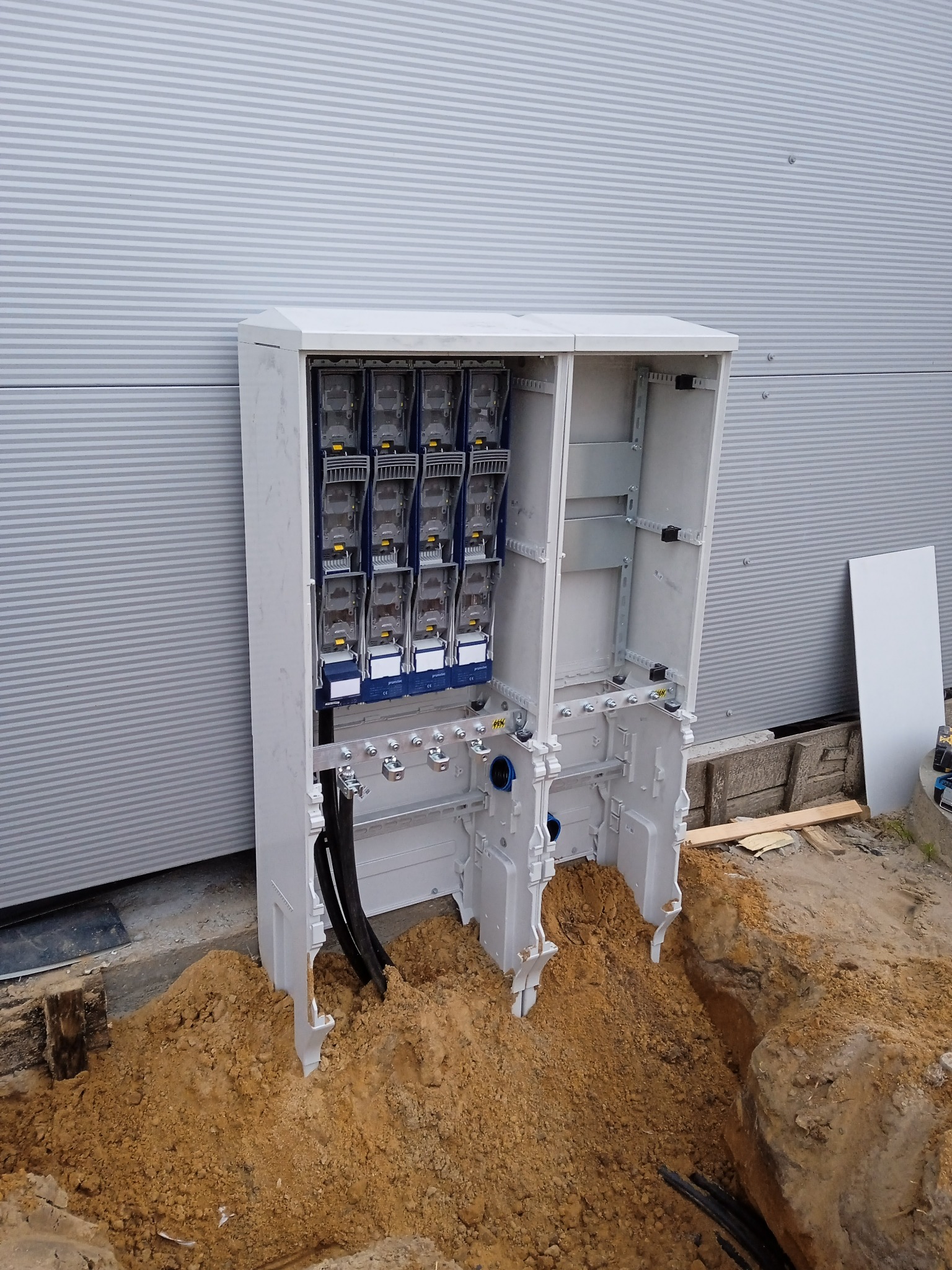 Otwarta, biała szafa elektryczna z zamontowanymi licznikami, umieszczona na zewnątrz budynku z szarej blachy falistej, w wykopie ziemnym. Widoczne kable.