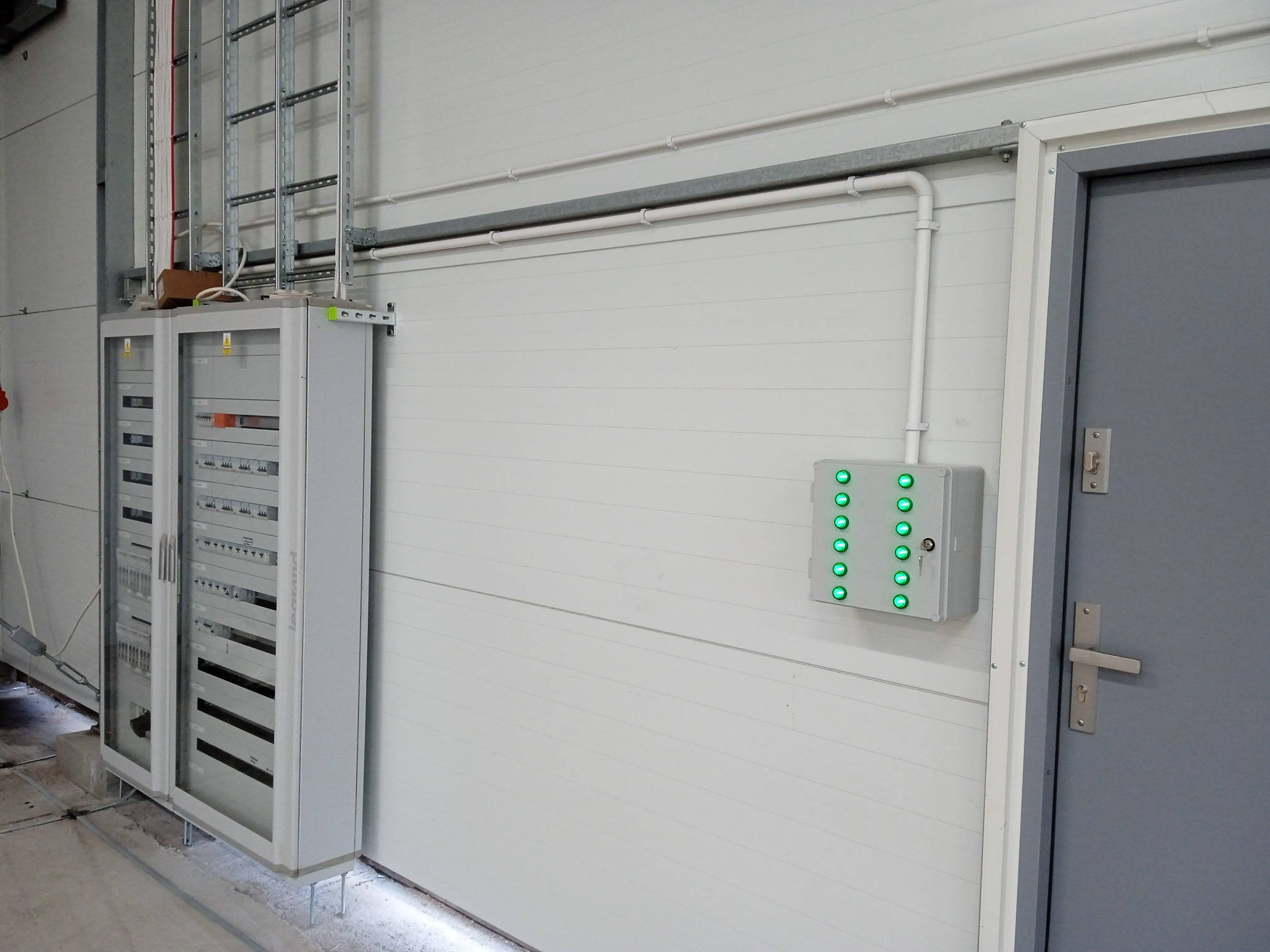 Szara szafa sterownicza z oszklonymi drzwiami i panel kontrolny z zielonymi diodami LED na białej ścianie w hali. Widoczne koryta kablowe pod sufitem i szare drzwi.