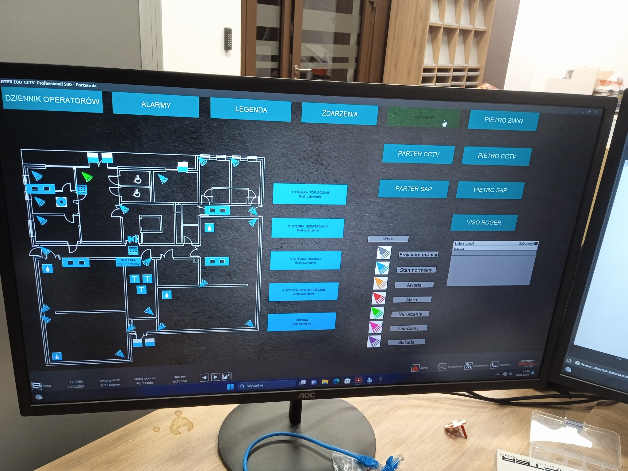 Ekran monitora z interfejsem systemu monitoringu: plan budynku z ikonami czujników, legendą i statusem. Widoczne okna z listą zdarzeń i stanem komunikacji.