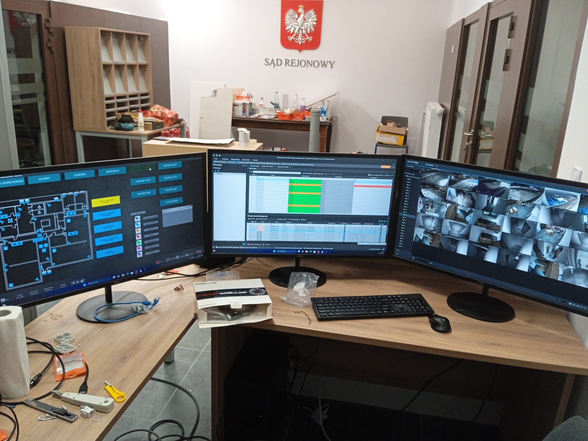 Stanowisko pracy z trzema monitorami prezentującymi schemat budynku, monitoring wizyjny i interfejs systemu. Na biurku narzędzia i akcesoria instalacyjne.