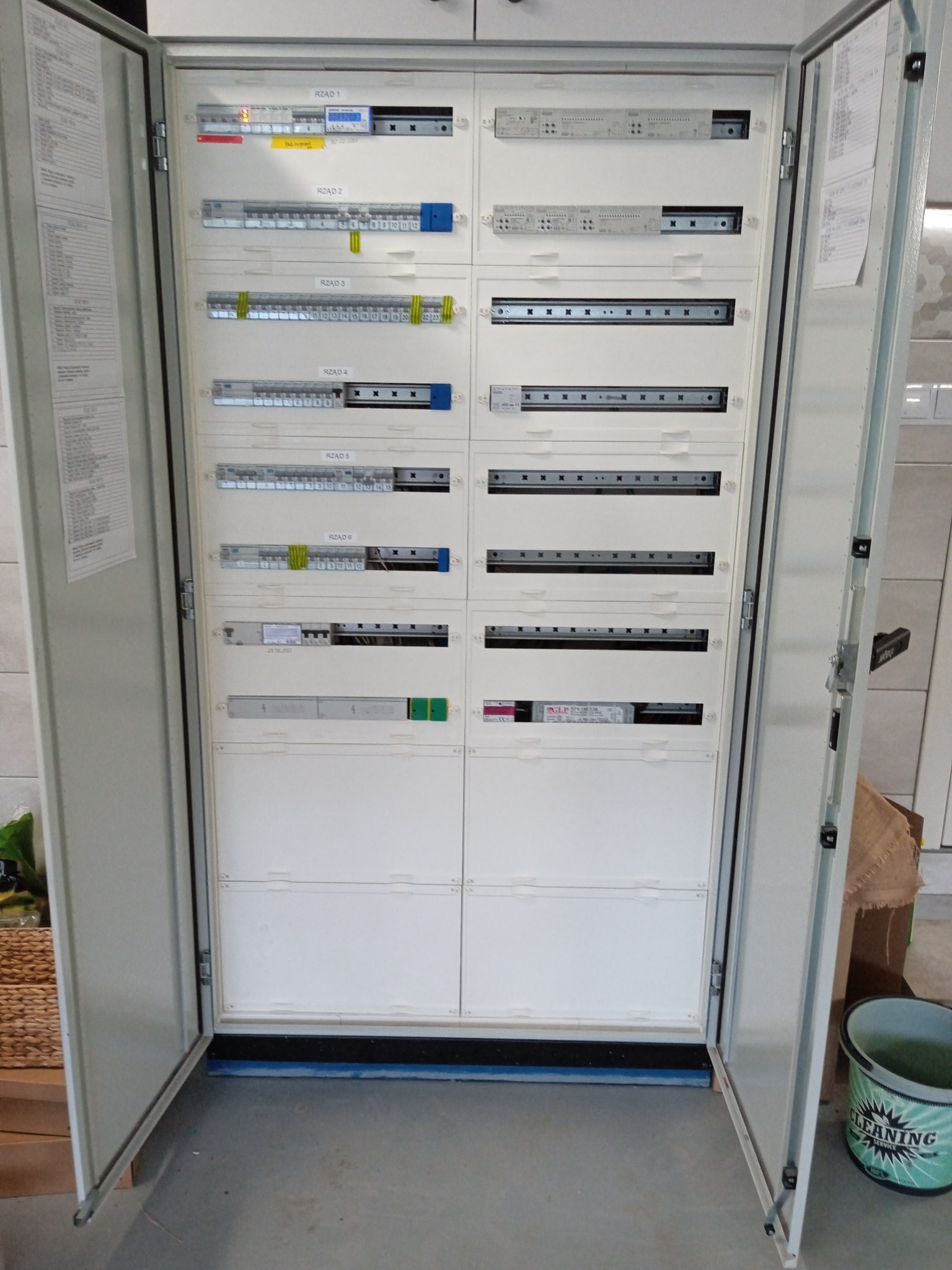 Otwarta szafa elektryczna z widocznymi rzędami zabezpieczeń i listwami, opisana jako RZĄD 1-6, w jasnym pomieszczeniu. Widoczne detale instalacji.