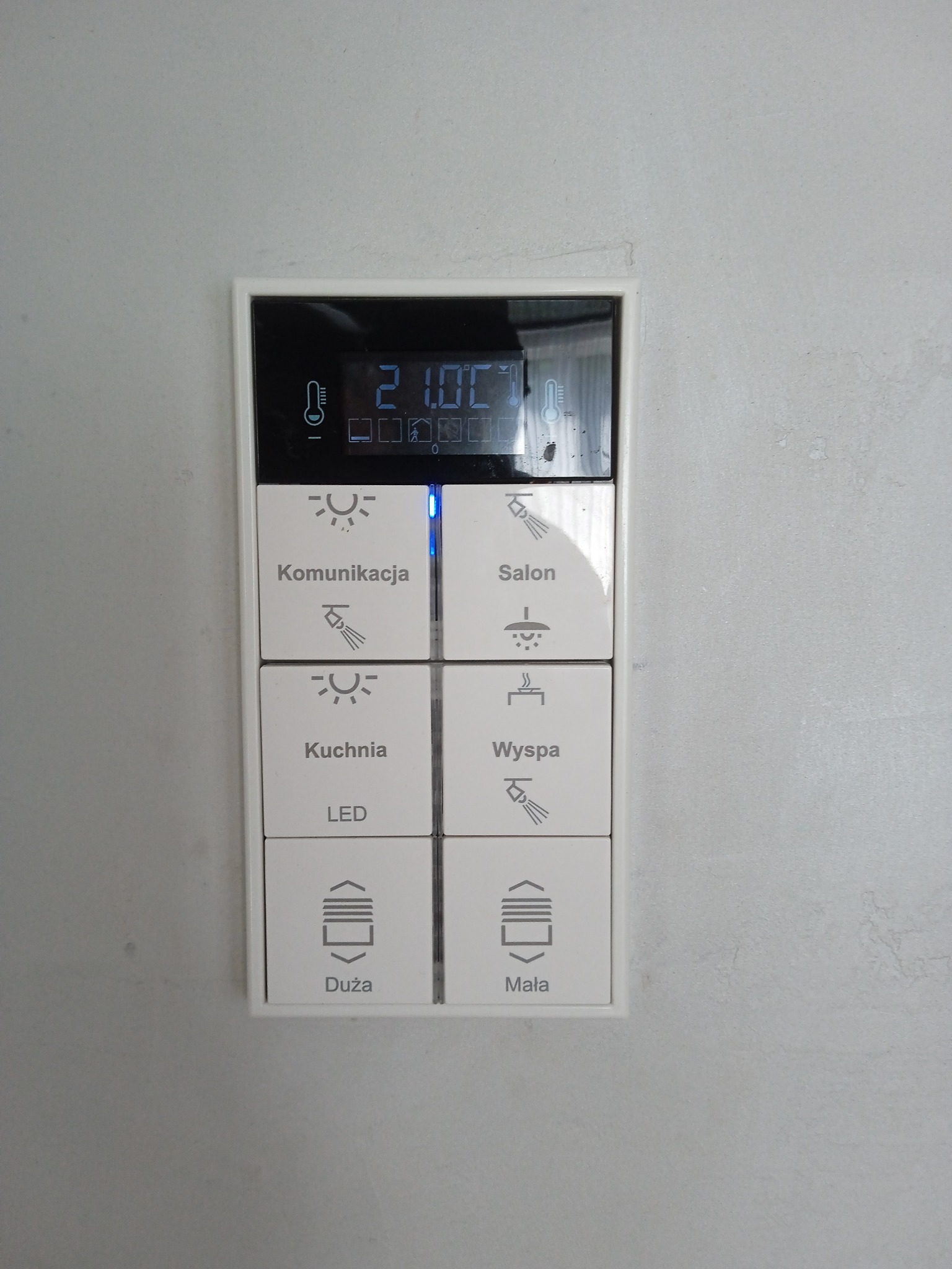 Panel sterowania inteligentnego domu z wyświetlaczem temperatury i ikonami oświetlenia dla różnych pomieszczeń, w tym kuchni, salonu i wyspy. Widoczne przyciski sterowania roletami.