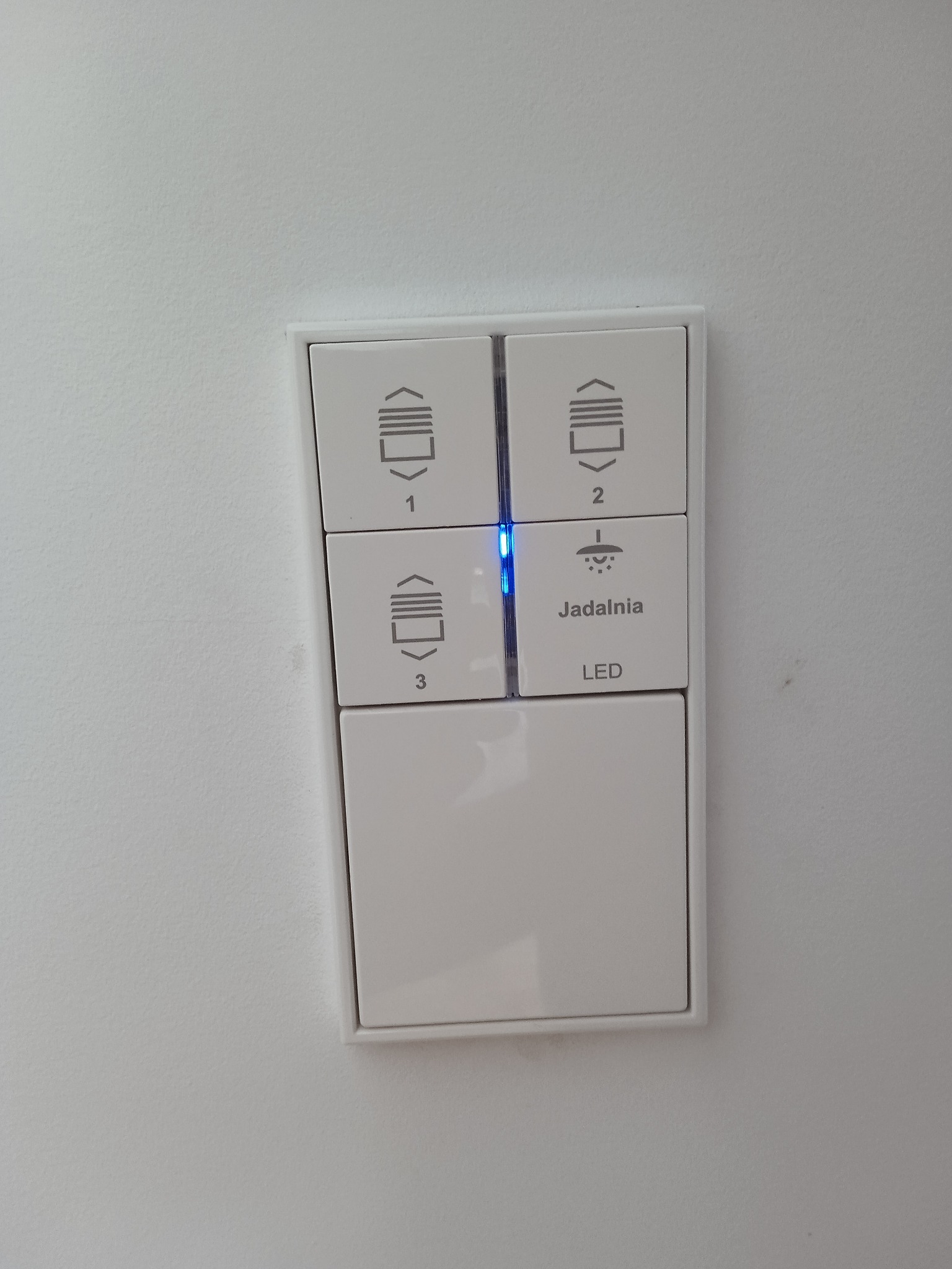 Panel sterowania inteligentnego domu z przyciskami do rolet (1, 2, 3) i oświetlenia (jadalnia), podświetlony diodą LED na białej ścianie.