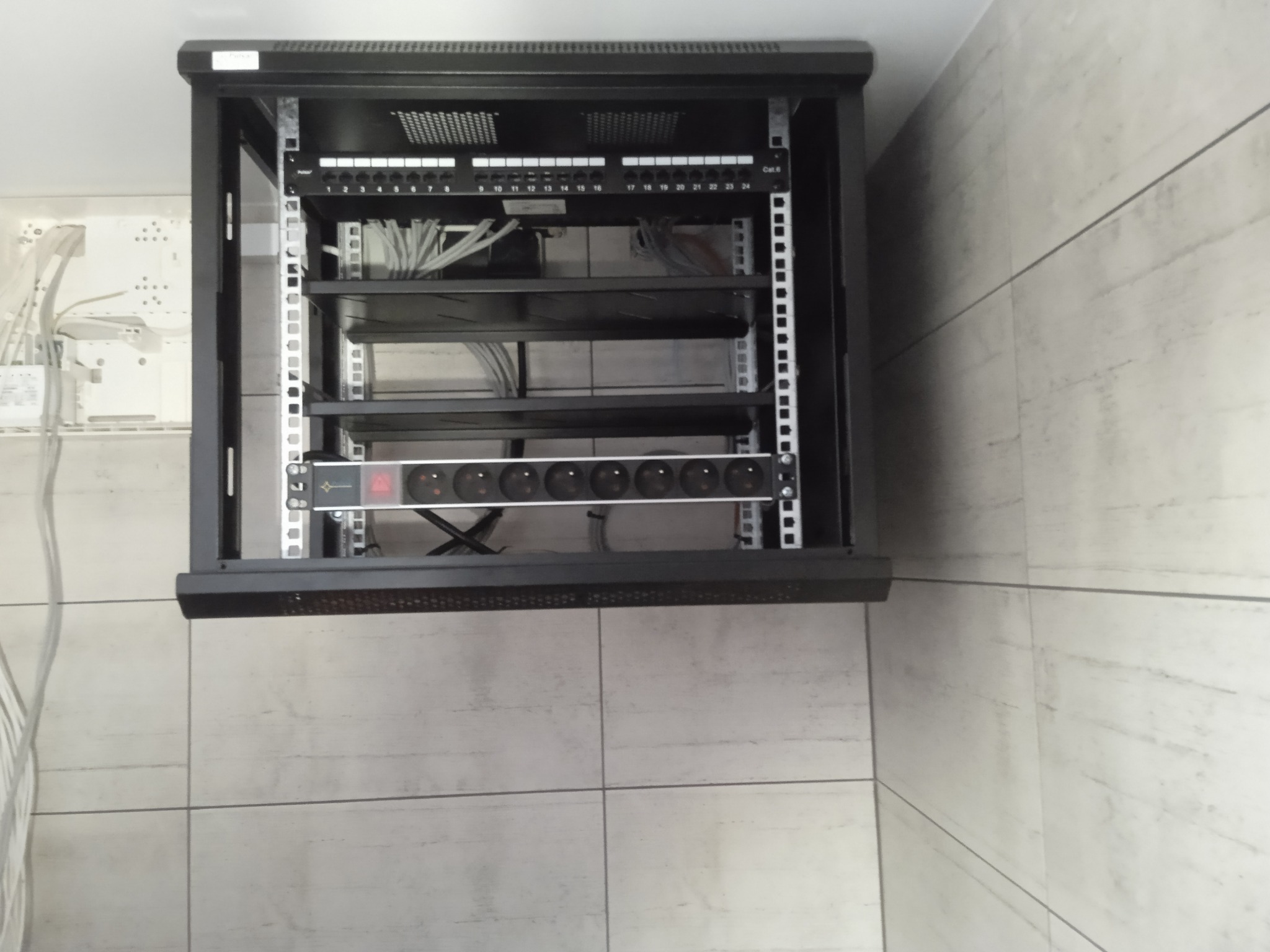 Zamontowana szafa rack z patch panelem, listwą zasilającą i okablowaniem w pomieszczeniu o jasnych, kwadratowych płytkach. Widoczna skrzynka z przewodami po lewej stronie.